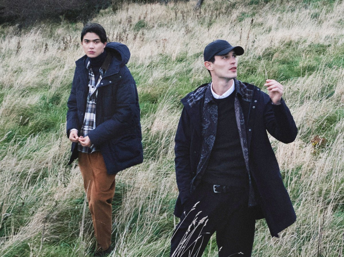 A man wearing a dark blue Rab rain jacket and a black backpack is outdoors, adjustss his jacket in a misty, mountainous landscape near a body of water.