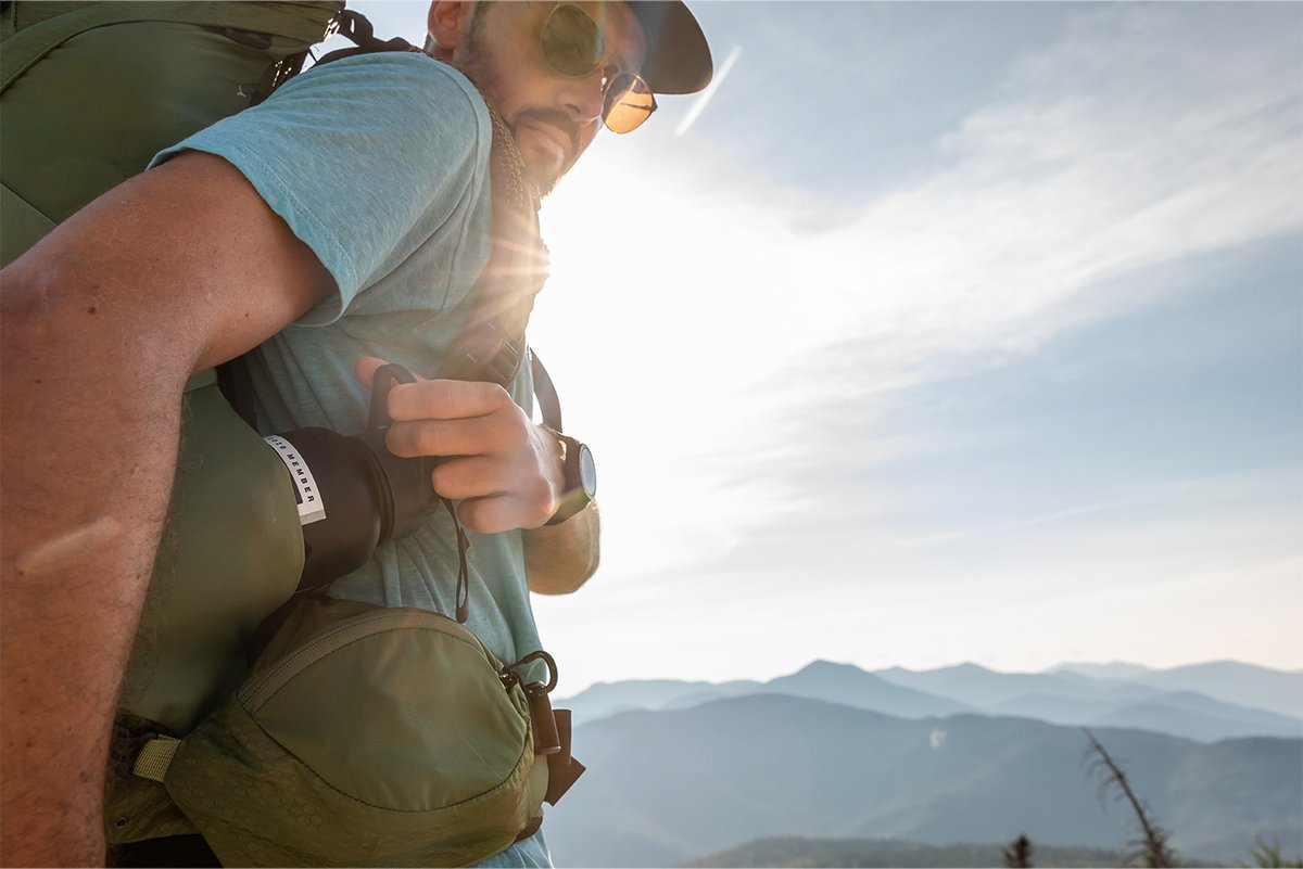 A close-up of a hiker in a blue shirt reaching for a water bottle in their green Osprey pack.