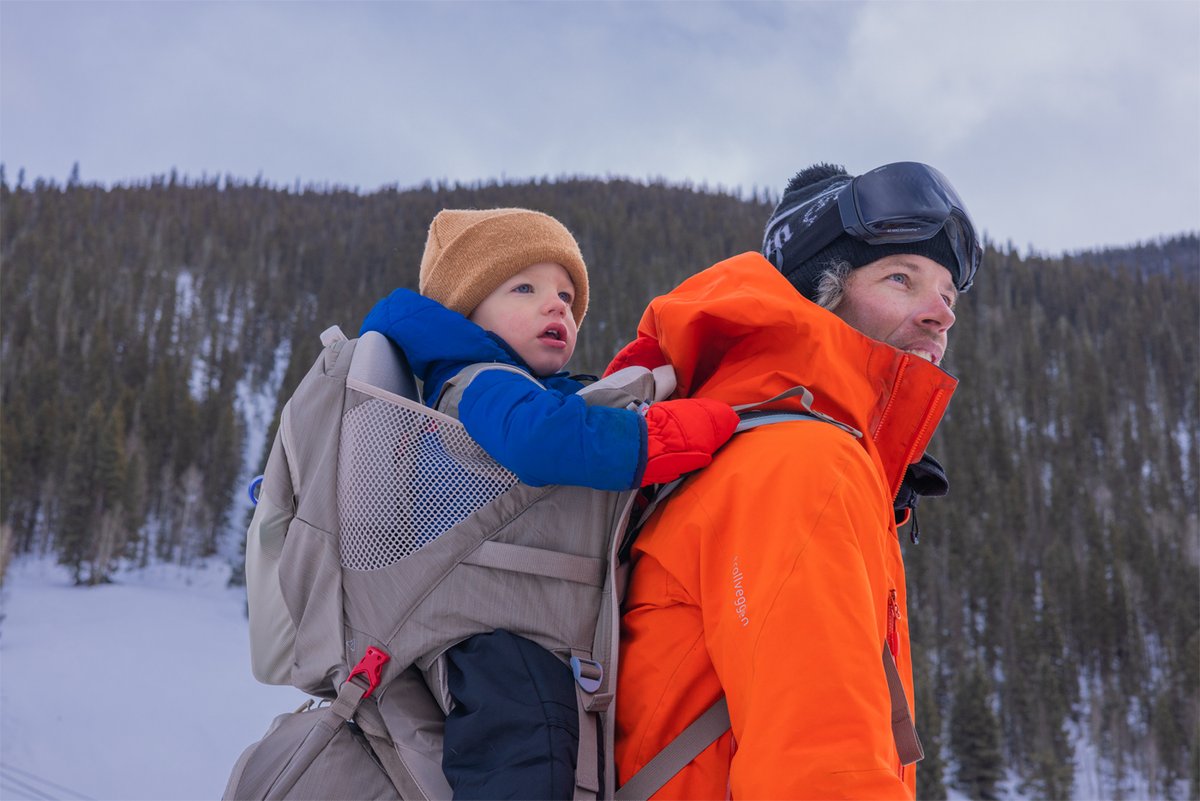 A man in an orange winter jacket and ski goggles carries a toddler in an Osprey child carrier while hiking in a snowy, mountainous forest.