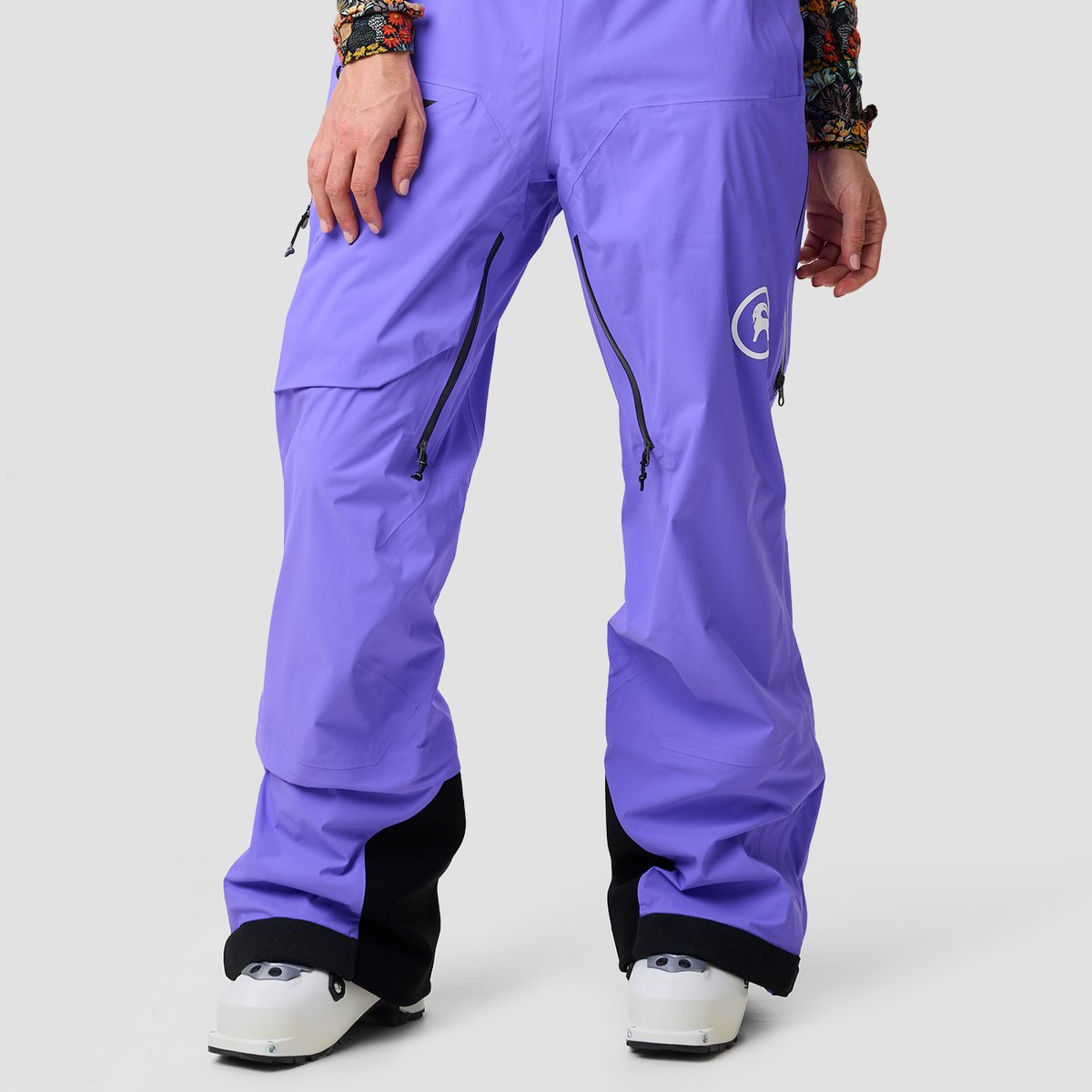 Close-up of a person's legs wearing lavender Cottonwoods ski pants, showing zippered thigh vents, a reflective brand logo on the right thigh, and reinforced black cuffs above white ski boots.