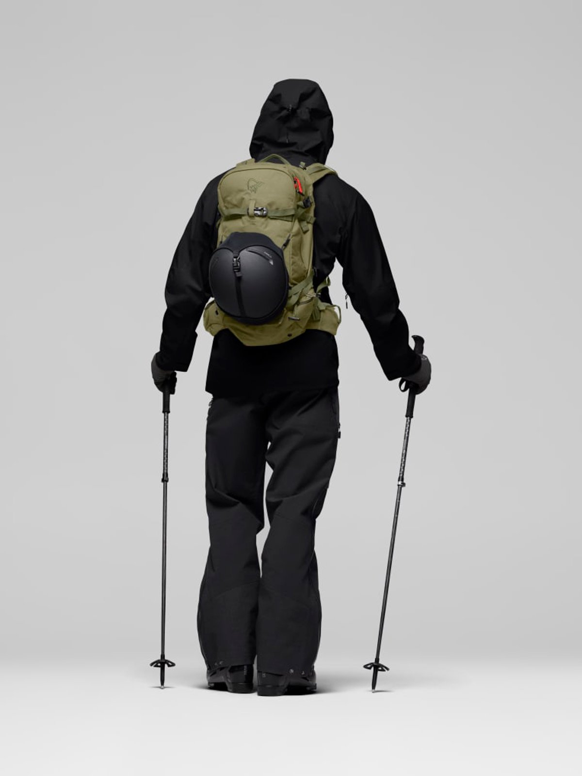 Rear view of a skier wearing a tan and black hiking backpack with attached helmet, black jacket, snow pants, and ski poles against a white background.