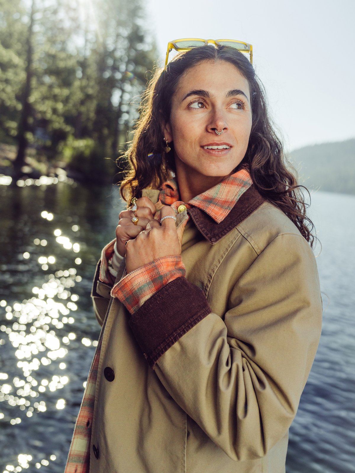 A woman with long wavy hair stands by a sunlit lake wearing sunglasses on her head, a tan coat with brown cuffs, and an orange plaid shirt, with trees and hills in the background.