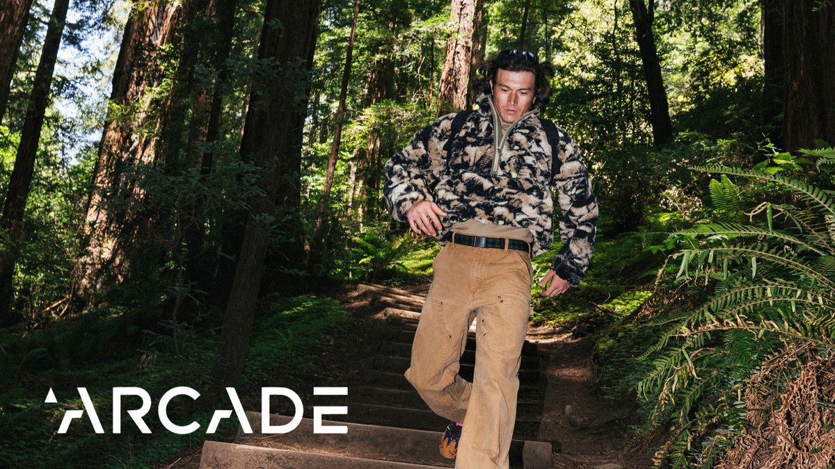 Man wearing patterned fleece jacket and tan work pants with Arcade belt standing on forest trail surrounded by tall trees and ferns.