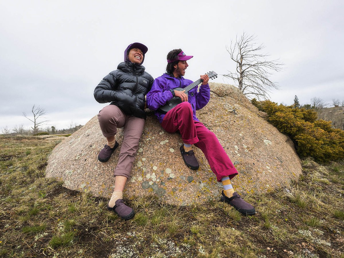Two people laughing on a granite boulder in a grassy meadow, one playing ukulele, both wearing Bedrock Mountain Clogs with socks under an overcast sky