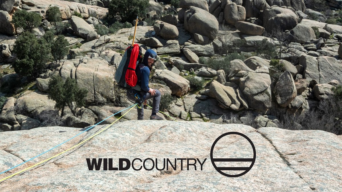 A rock climber in blue shirt and helmet rappels down a granite boulder field while secured by a blue rope, with a red coiled rope on their backpack.