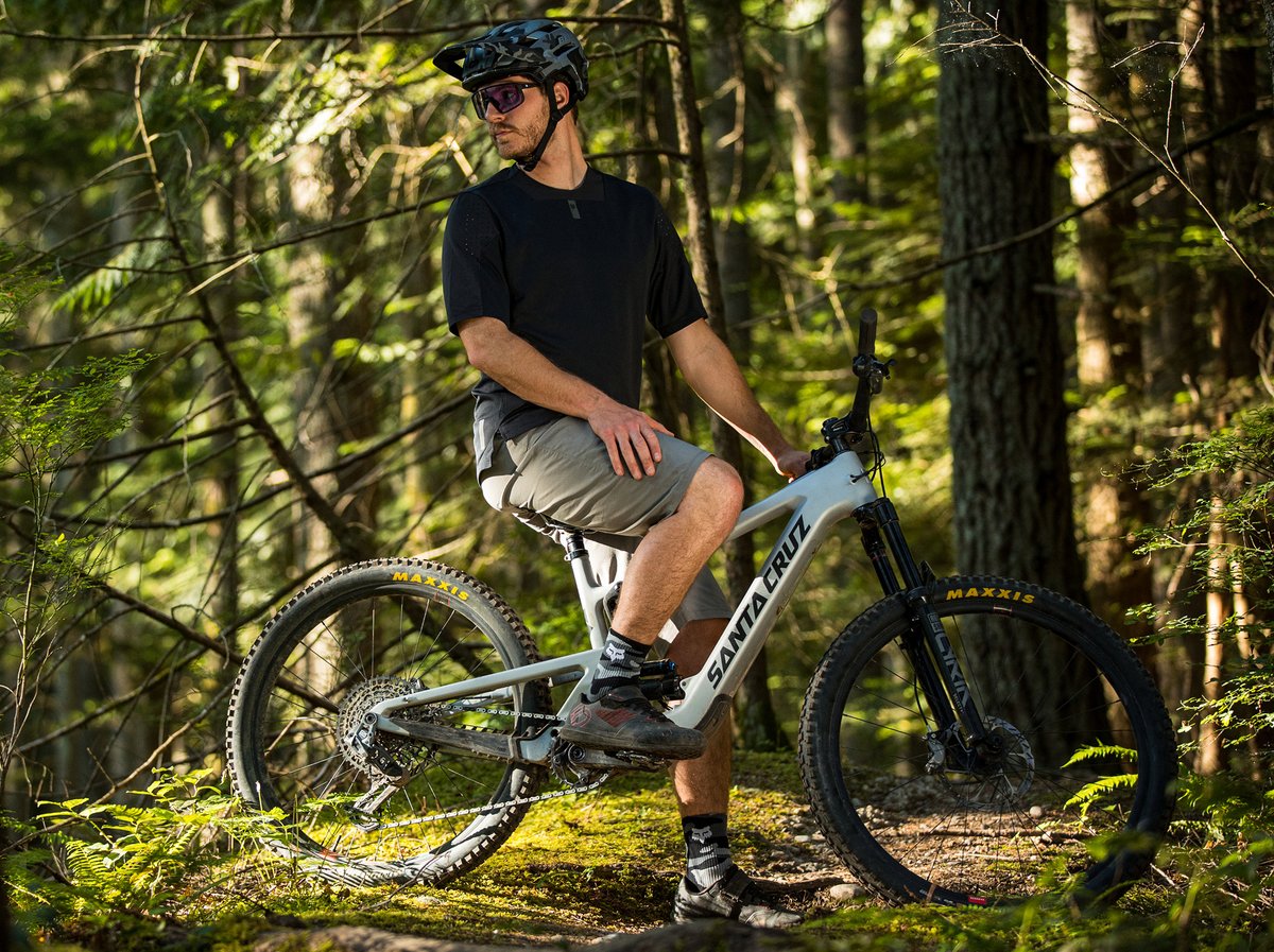 A man wearing a helmet and casual biking clothes sits on a Santa Cruz  mountain bike in a sunlit forest.