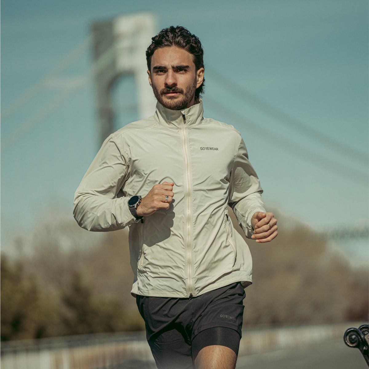 A person wearing a light-colored GOREWEAR jacket and black shorts is running outdoors with a bridge in the background.