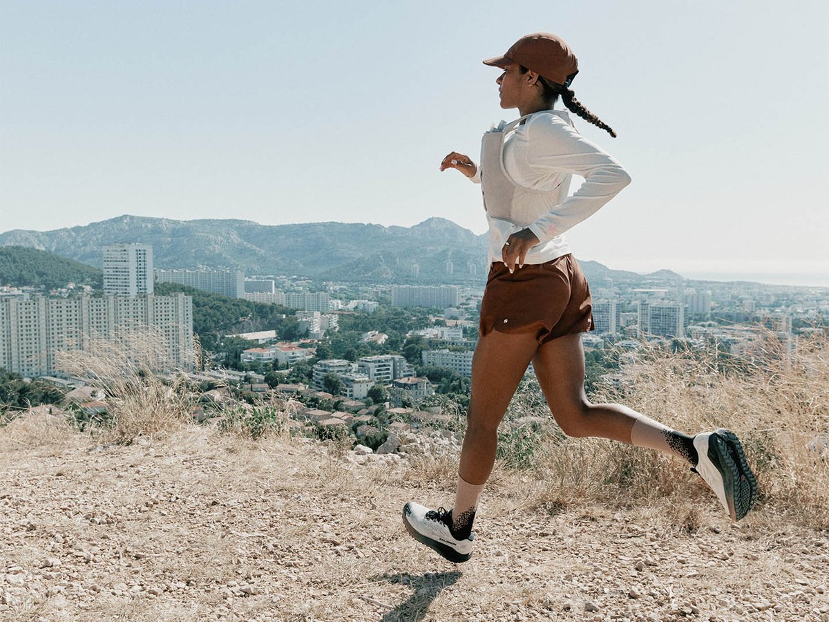 Female runner in profile on dry gravel hillside above city skyline, Salomon Aero Blaze 4 GRVL GTX