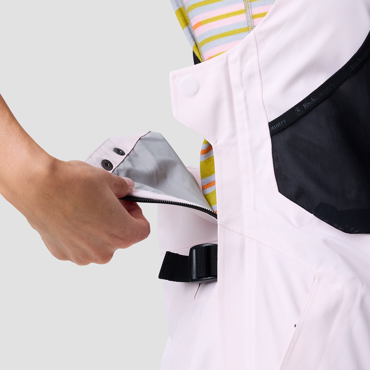 Close-up of a hand unsnapping the drop-seat flap on light pink Cottonwoods bibs, showing the two-snap closure over a zipper, mesh ventilation panel, and adjustable back-waist strap with a black cam buckle.