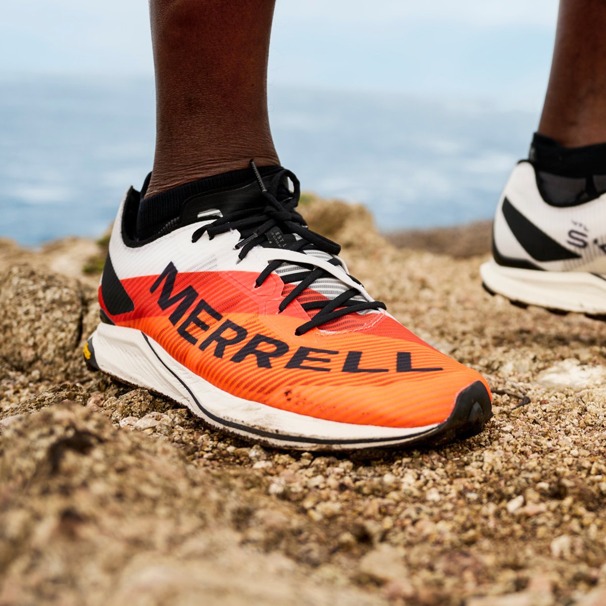 A person wearing bright orange and white Merrell MTL running shoes with compact Vibram soles stands on rocky terrain. 