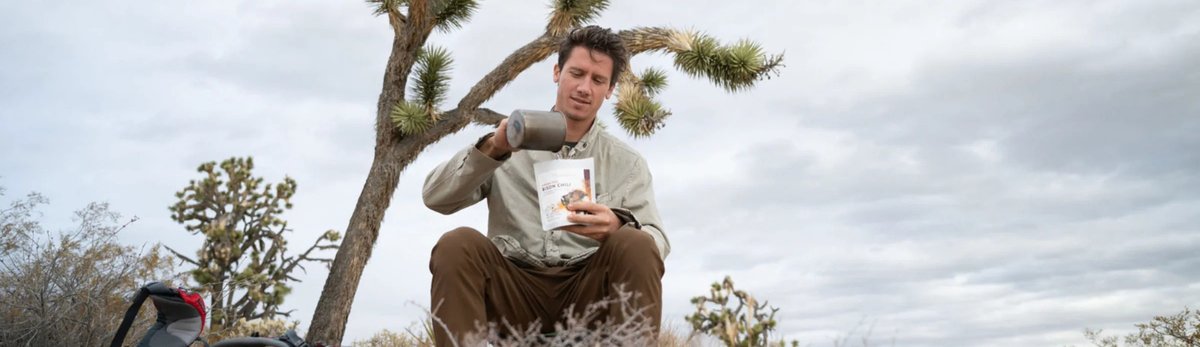 Backpacker sits in a desert landscape beneath a Joshua tree, pouring hot water into a meal pouch with a backpack beside him.