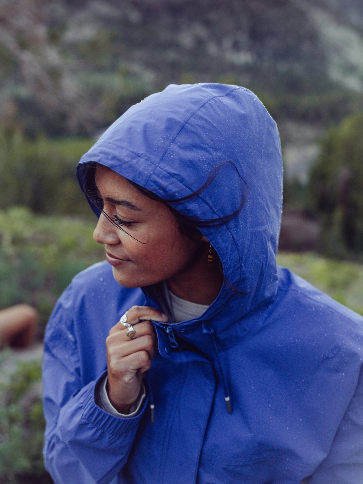 A woman in a blue hooded rain jacket stands outdoors in a natural, mountainous area.