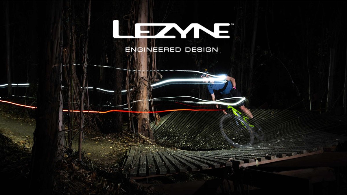 A mountain biker rides through a dark forest trail at night, illuminated by LED bike lights, with the Lezyne "Engineered Design" logo displayed above.