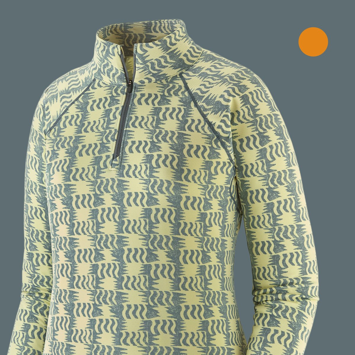 Close-up of a pale yellow and grey zigzag-print quarter-zip baselayer top, showing the collar and zipper pull, on a slate-grey background with an orange swatch.