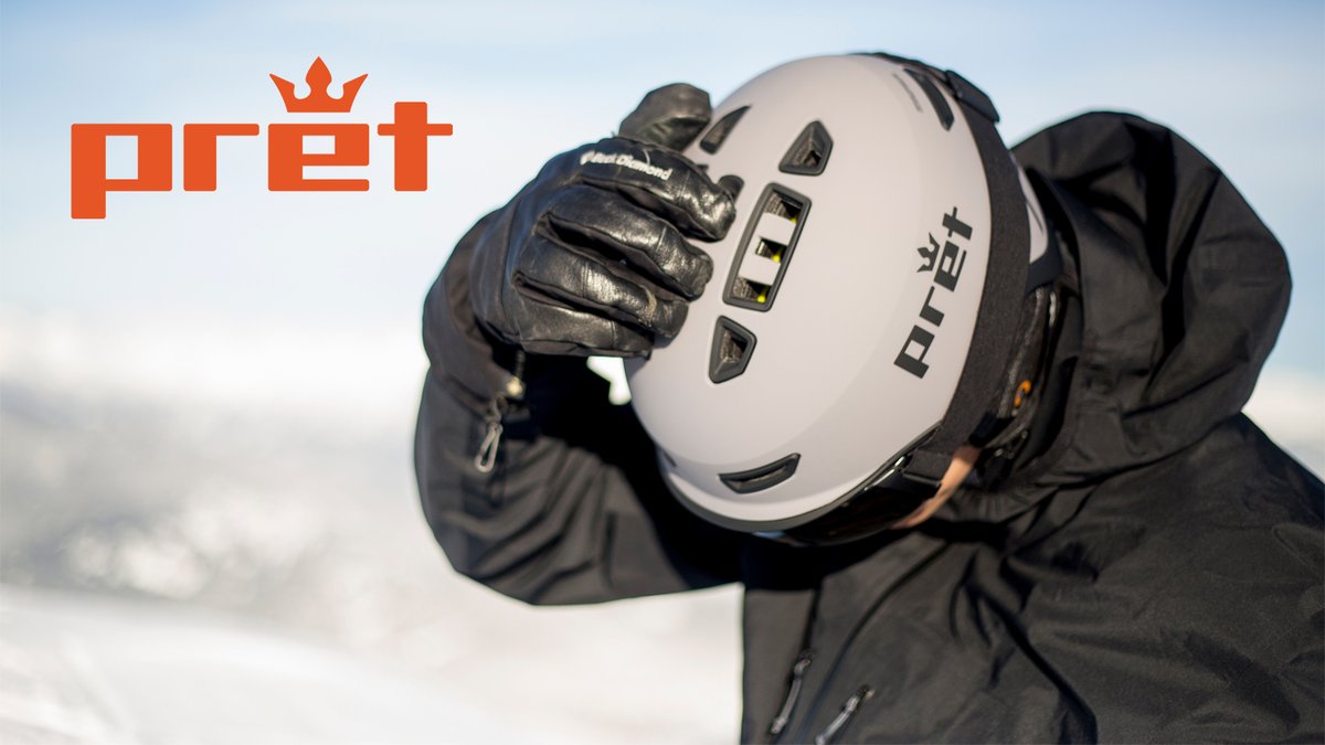 Person wearing a white Pret ski helmet with black gloves holds the helmet up against a snowy mountain backdrop.