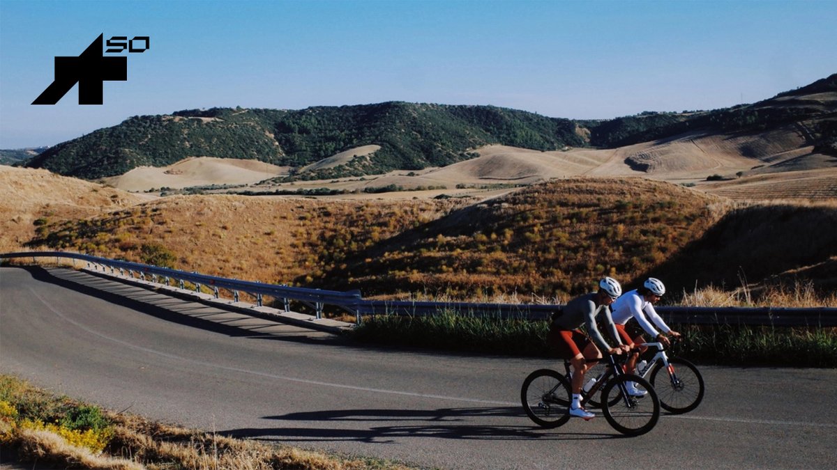 Two cyclists in Assos gear ride along a winding mountain road through a hilly, dry landscape.