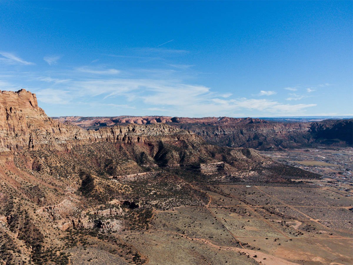  Aerial view of a broad red sandstone mesa with layered cliff faces dropping to a wide desert valley, with a small community visible at the far right edge.