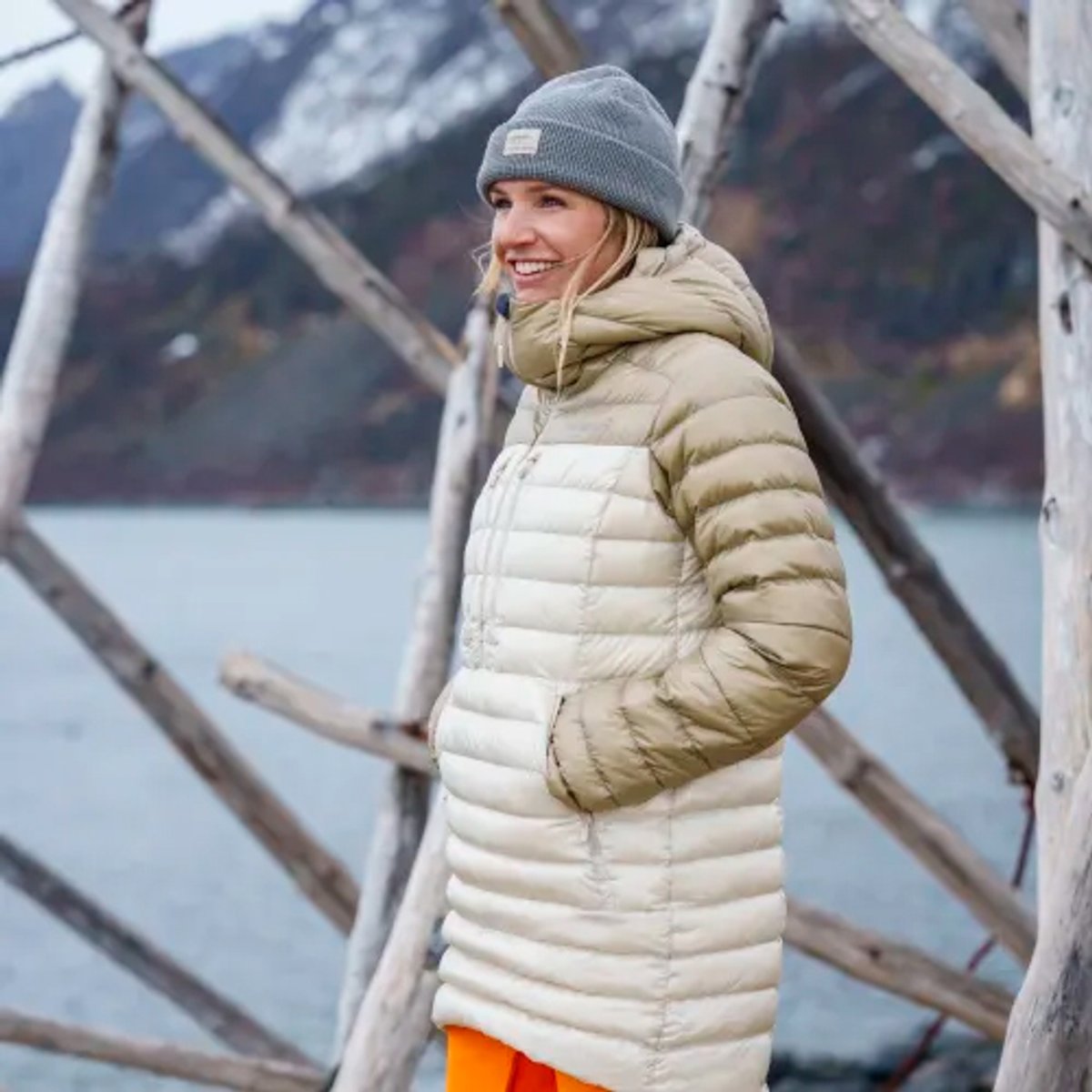 Woman wearing a beige and tan puffer jacket with gray beanie standing outdoors by a lake in winter.