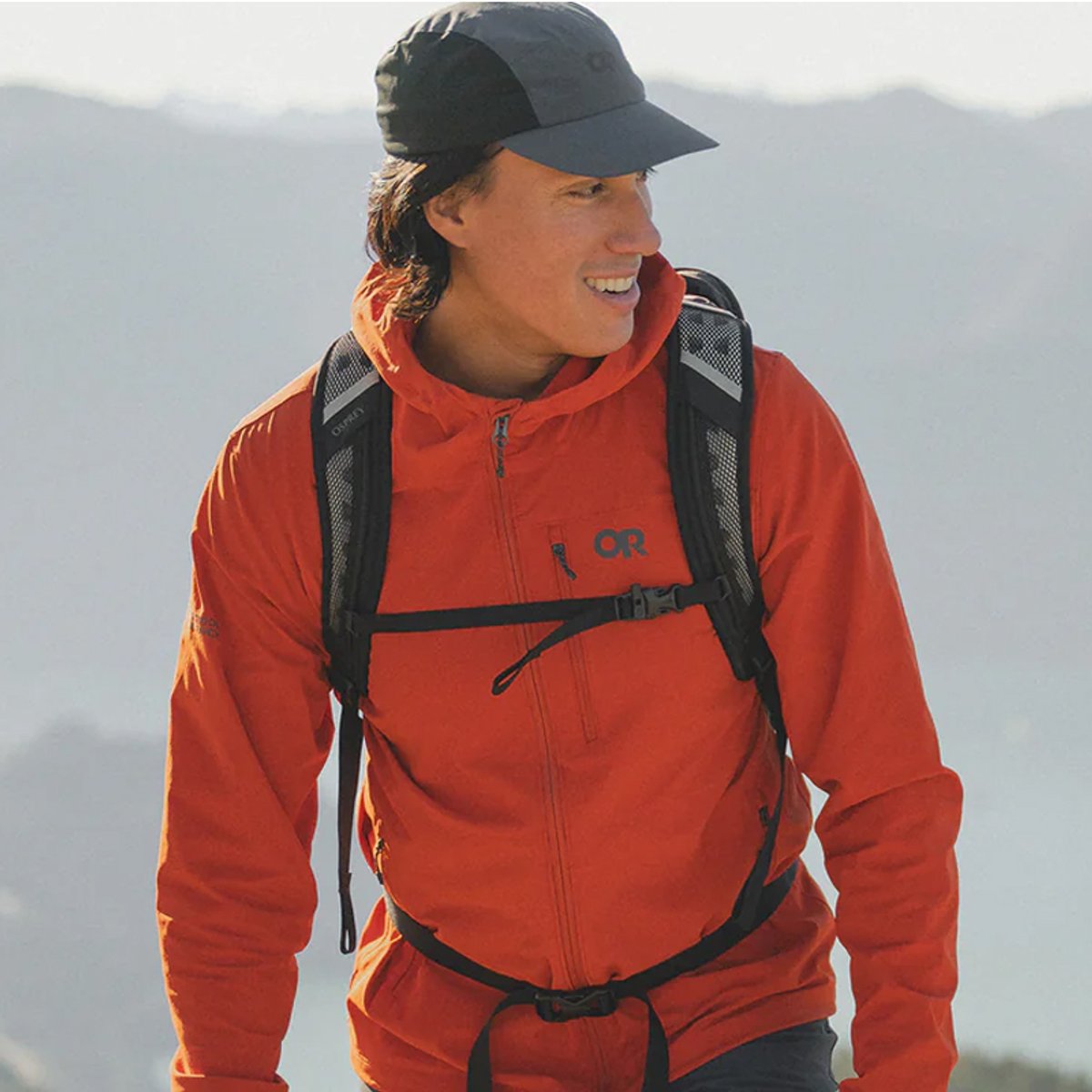 A man wears a Ferrosi hoodie, hat, and backpack, walking against the backdrop of mountains.