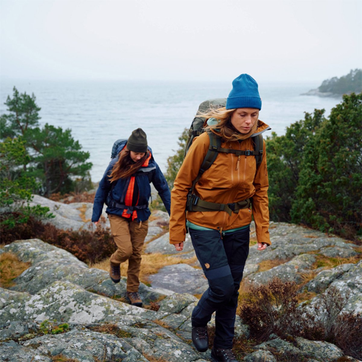 Two hikers wearing backpacks and winter hats trek across rocky coastal terrain with a lake or ocean visible in the misty background.