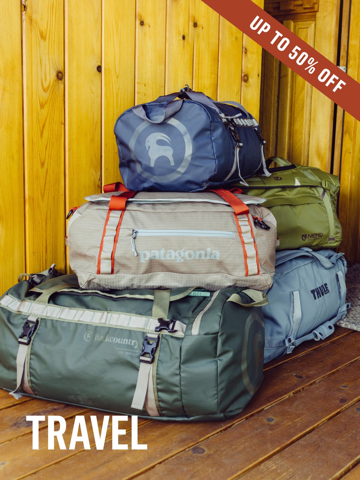 A pile of travel duffels — Backcountry, Patagonia, Thule, and NEMO — stacked against a wood wall. Up to 50% off travel gear.