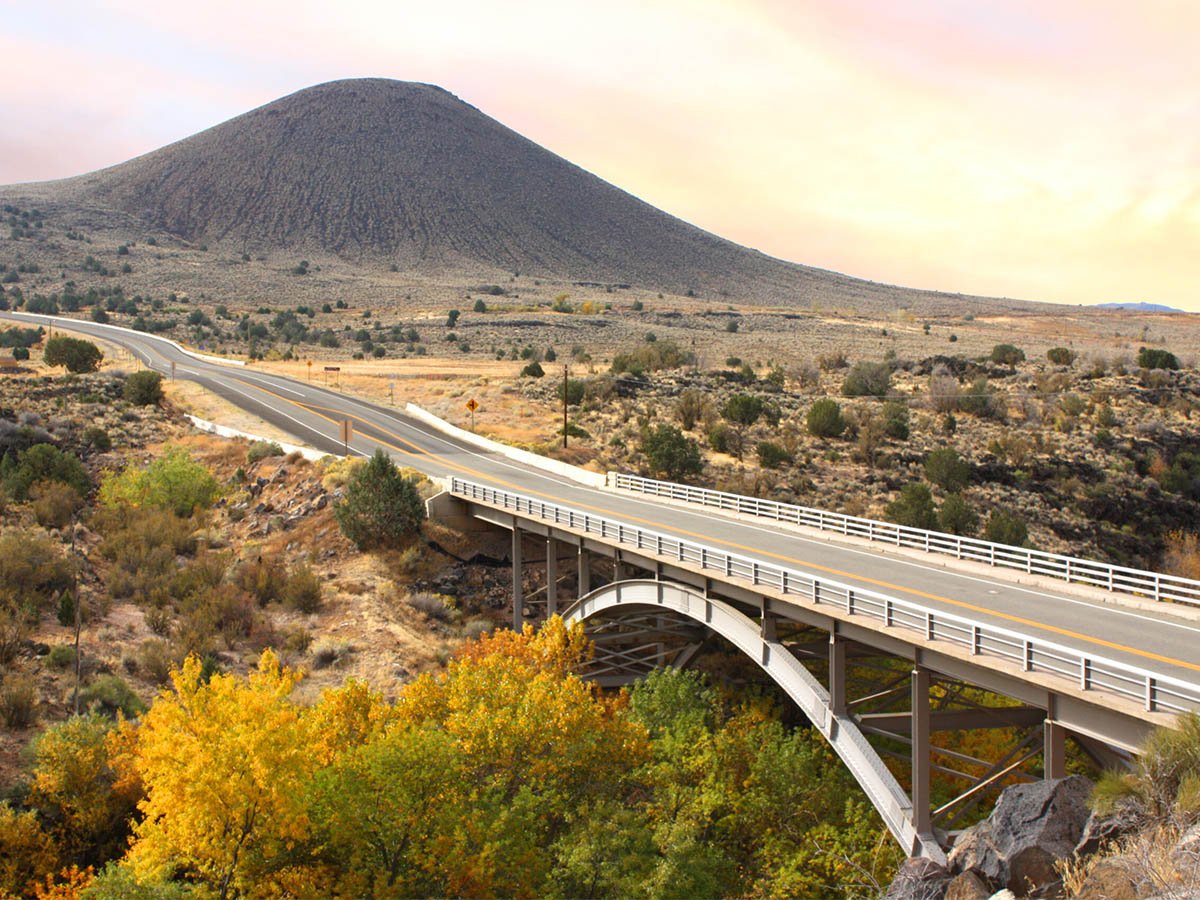 Two-lane highway bridge spanning a canyon above fall-colored cottonwoods, with a dark volcanic cinder cone rising from the desert scrub in the background under a warm sunset sky.