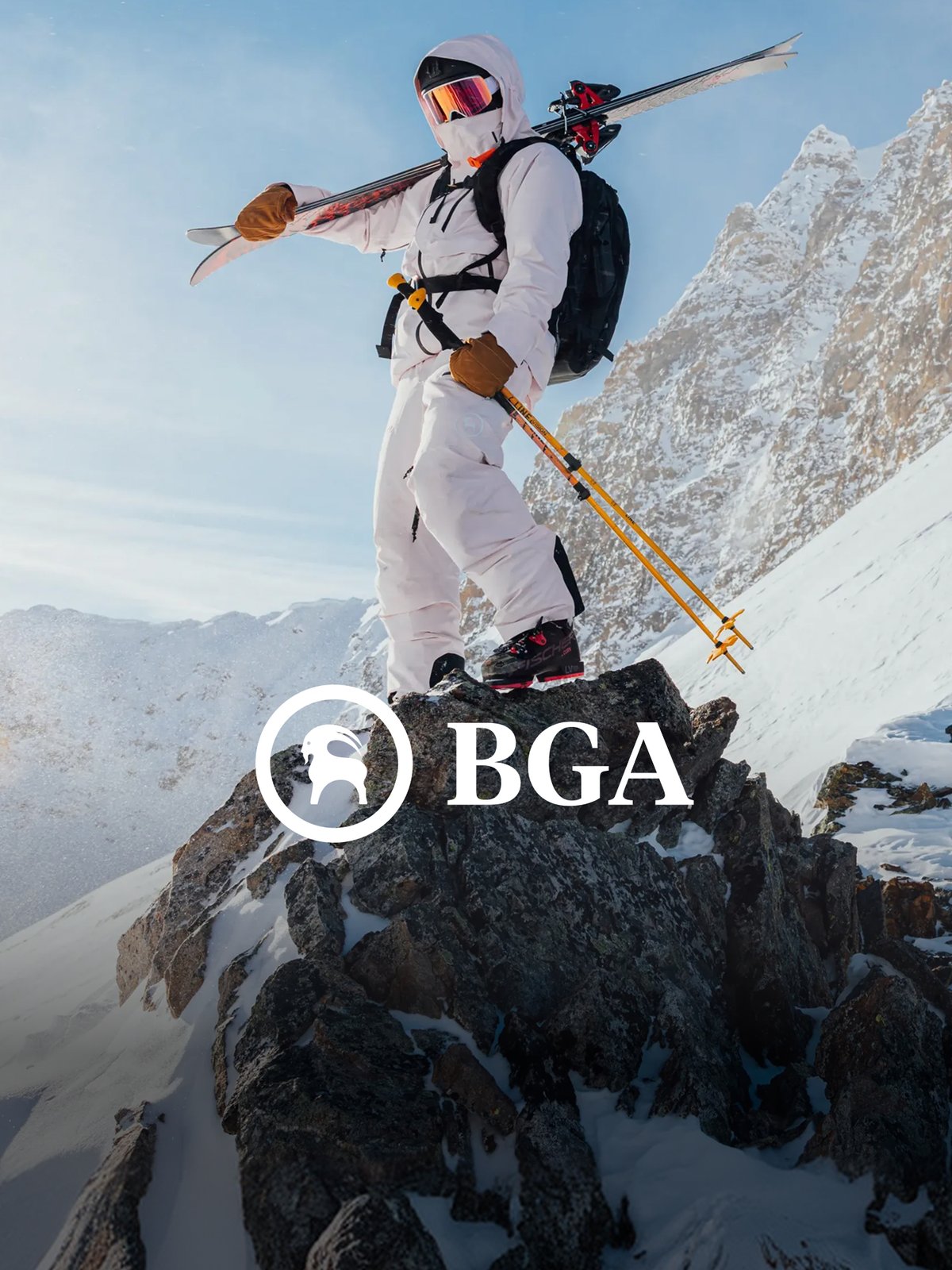 Skier in a light pink BGA outerwear kit suit standing on a rocky alpine summit with skis over one shoulder, featuring the BGA logo.