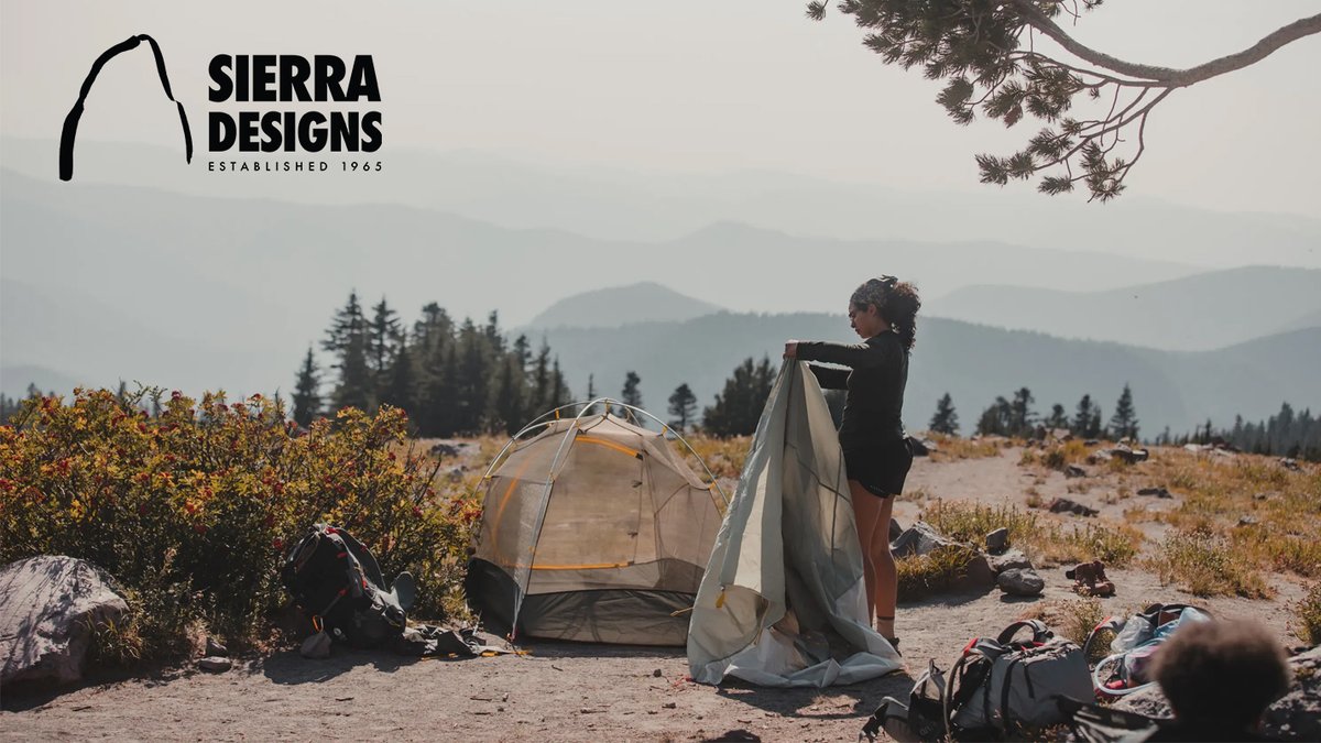 Woman setting up a lightweight Sierra Designs camping tent in a mountainous landscape with coniferous forest and golden shrubland during a backpacking trip.