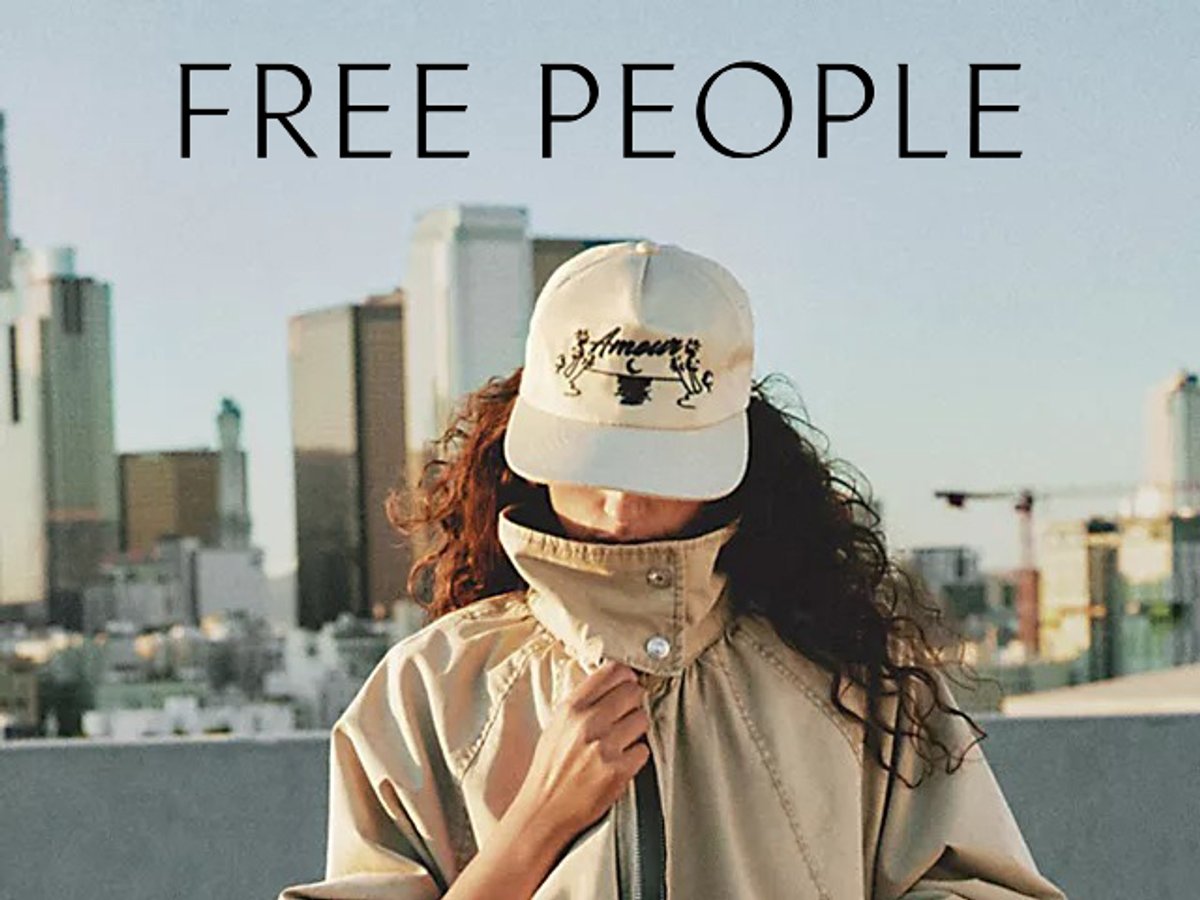 A woman wearing an "Amour" baseball cap and a tan jacket with a city skyline in the background; "FREE PEOPLE" text is at the top