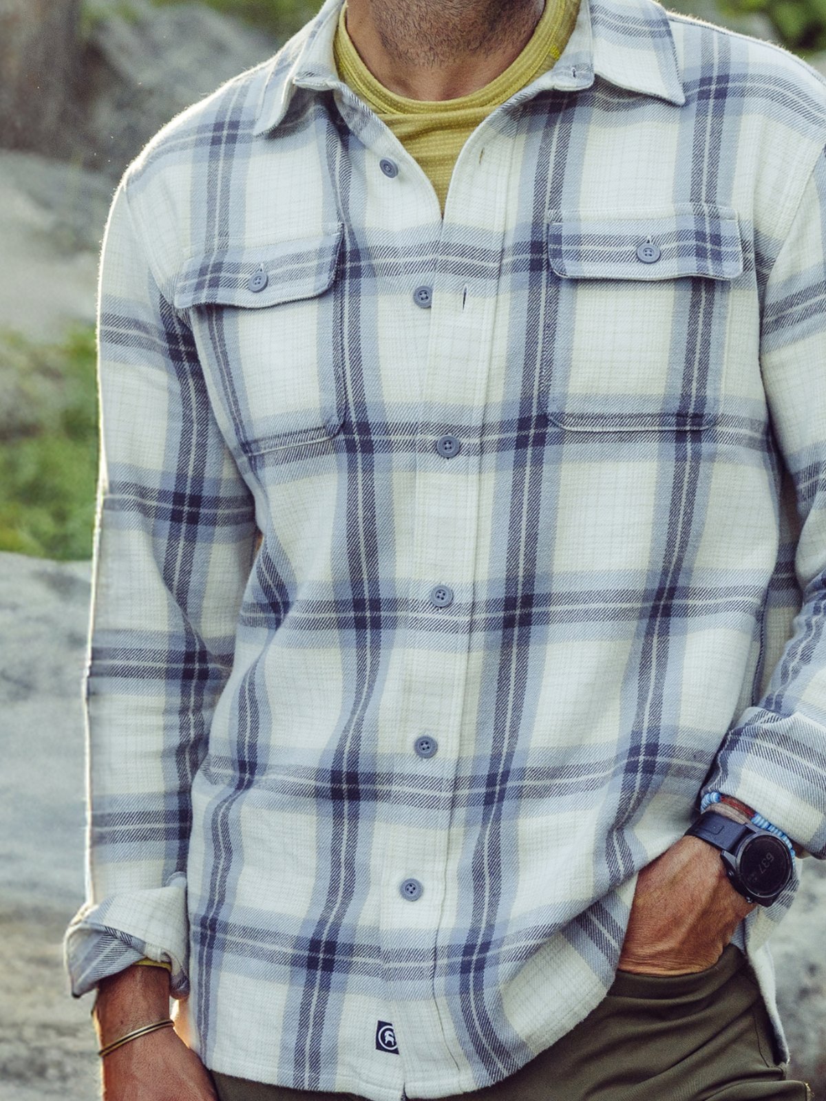 A person stands outdoors wearing a light-colored plaid button-up shirt over a yellow crew-neck top, with one hand in the pocket of olive green pants and a wristwatch visible.