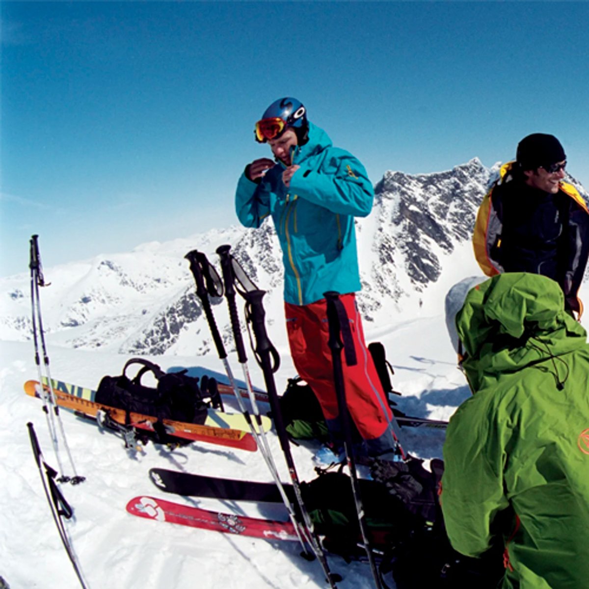Group of skiers in colorful winter gear standing on snowy mountain slope with ski equipment and alpine peaks in background.