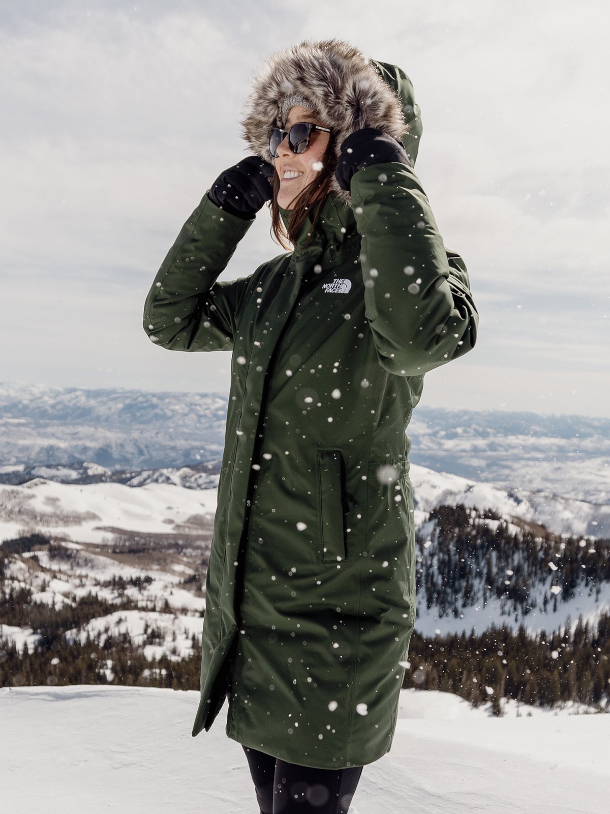 A in a green The North Face parka with a fur-lined hood stands on a snowy mountain.