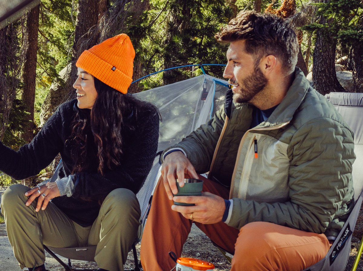 A woman and a man dressed in fleece jackets and vest share coffee at camp. Overlaid text reads, 