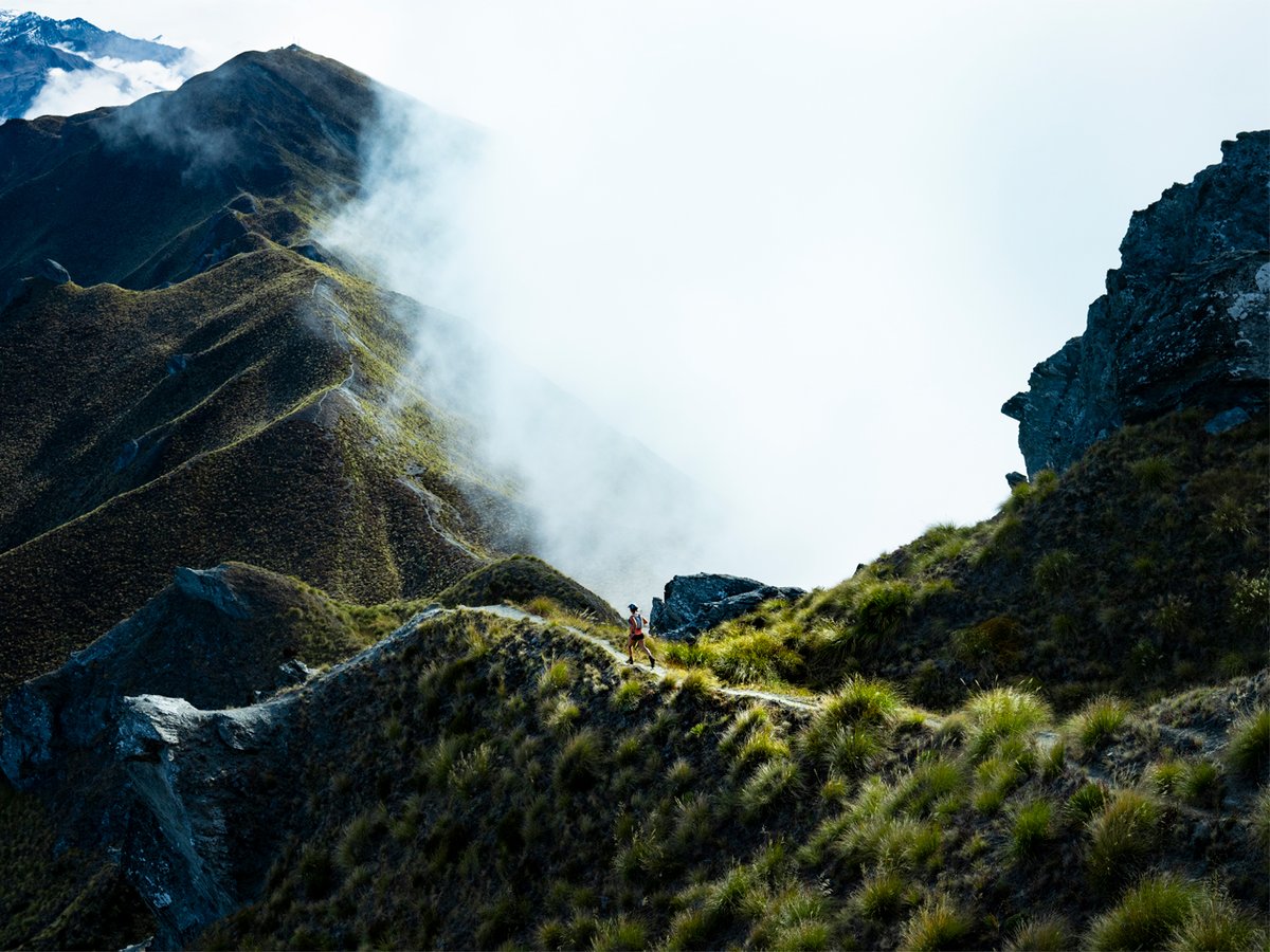 A woman runs in the distance, amid a mountain landscape filled with fog.