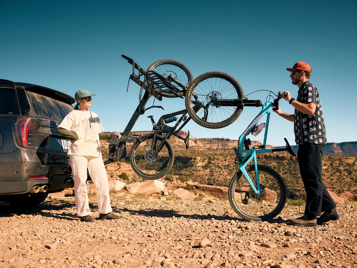 A man and a woman stand next to an SUV parked in a desert landscape. The man is holding a teal Santa Cruz full-suspension mountain bike vertically, lifting the front wheel to load it onto a hitch-mounted vertical bike rack on the back of the SUV, which already holds a black mountain bike.