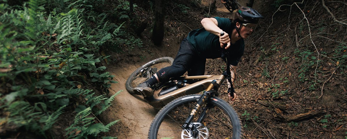 Mountain biker wearing MTB helmet and jersey railing tight singletrack turn on full-suspension mountain bike through dense forest trail.