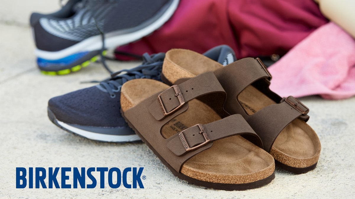 A pair of brown two-strap Birkenstock sandals sits on a concrete surface next to a pair of athletic sneakers and a pink towel.