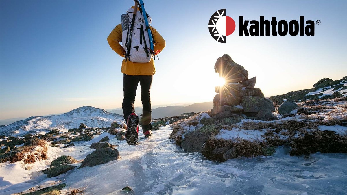 Hiker wearing a Kahtoola backpack and yellow jacket ascending a snowy mountain ridge at sunrise with rocky terrain and distant peaks visible in the background.