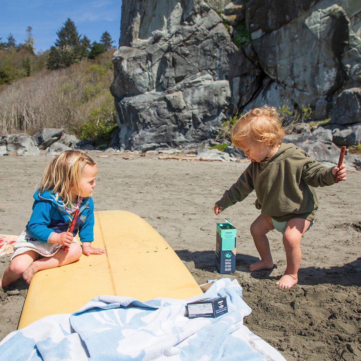 Two young children playing on a sandy beach next to a yellow surfboard with rocky cliffs in the background on a sunny day.