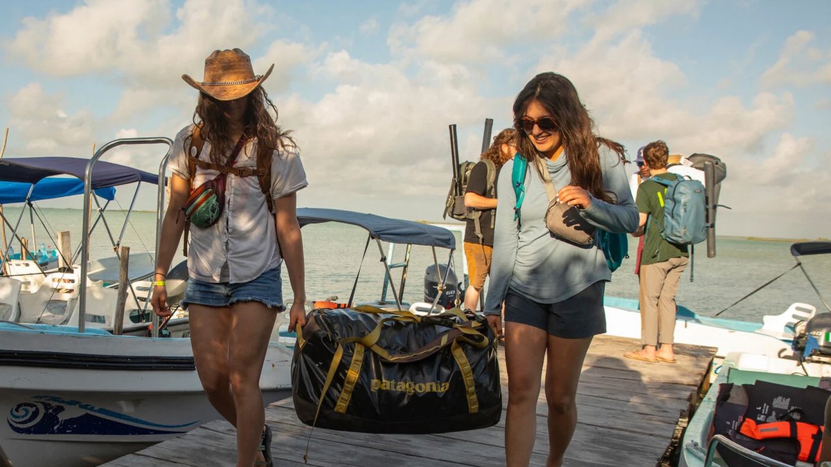 Two women carry each end of a Patagonia Black Hole duffel, while men stand behind them on the pier wearing Black Hole backpacks.