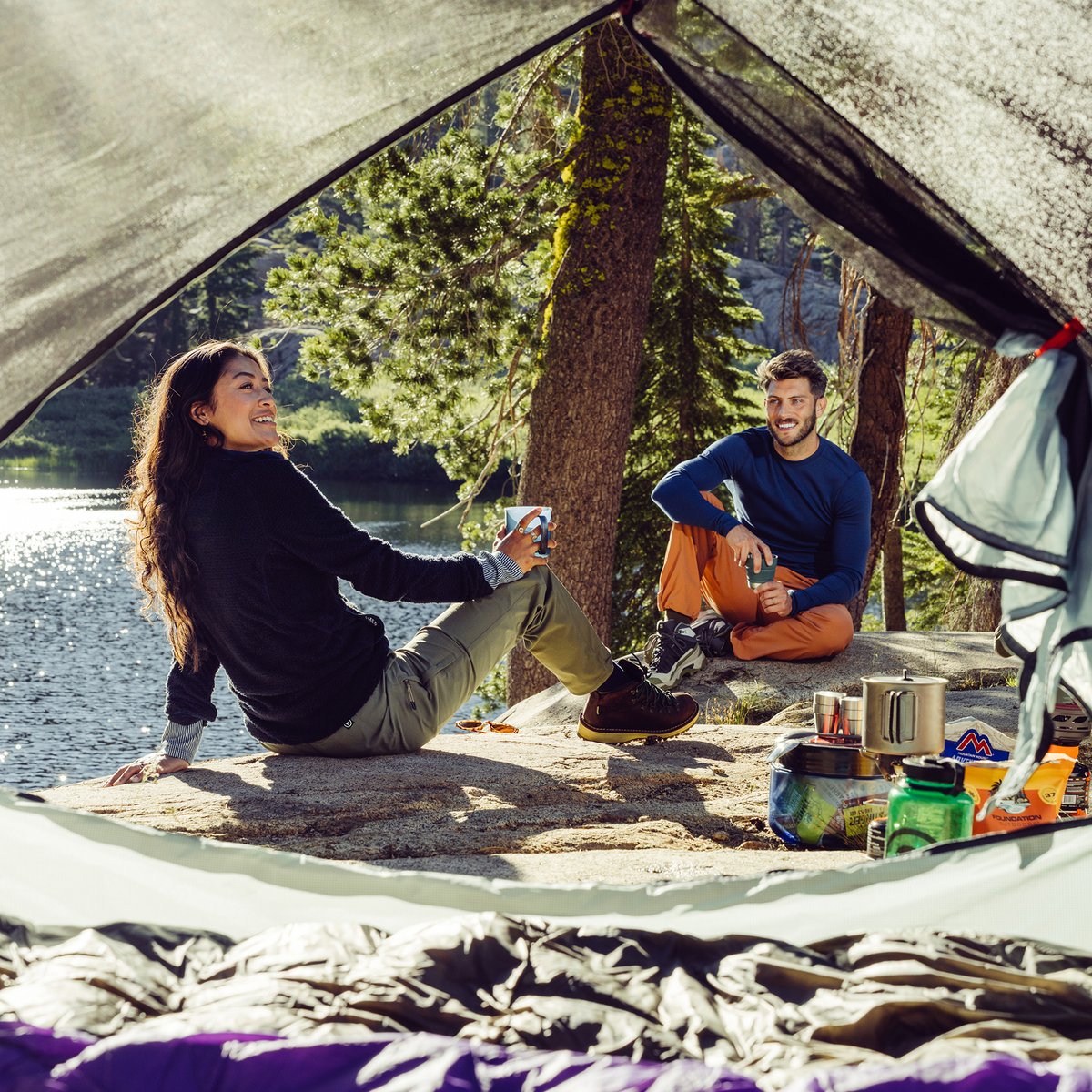 Two campers eating freeze-dried meals on volcanic terrain, laughing beside a pink tent and camp stove at dusk.