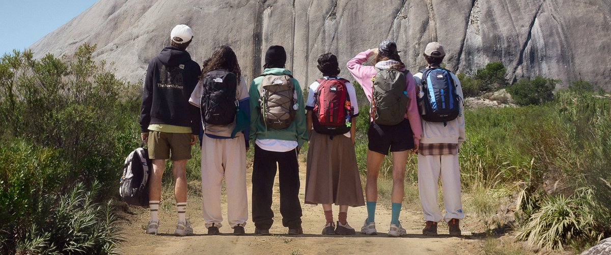 Six people wearing a variety of The North Face backpacks stand facing a large granite rock face in a desert landscape.
