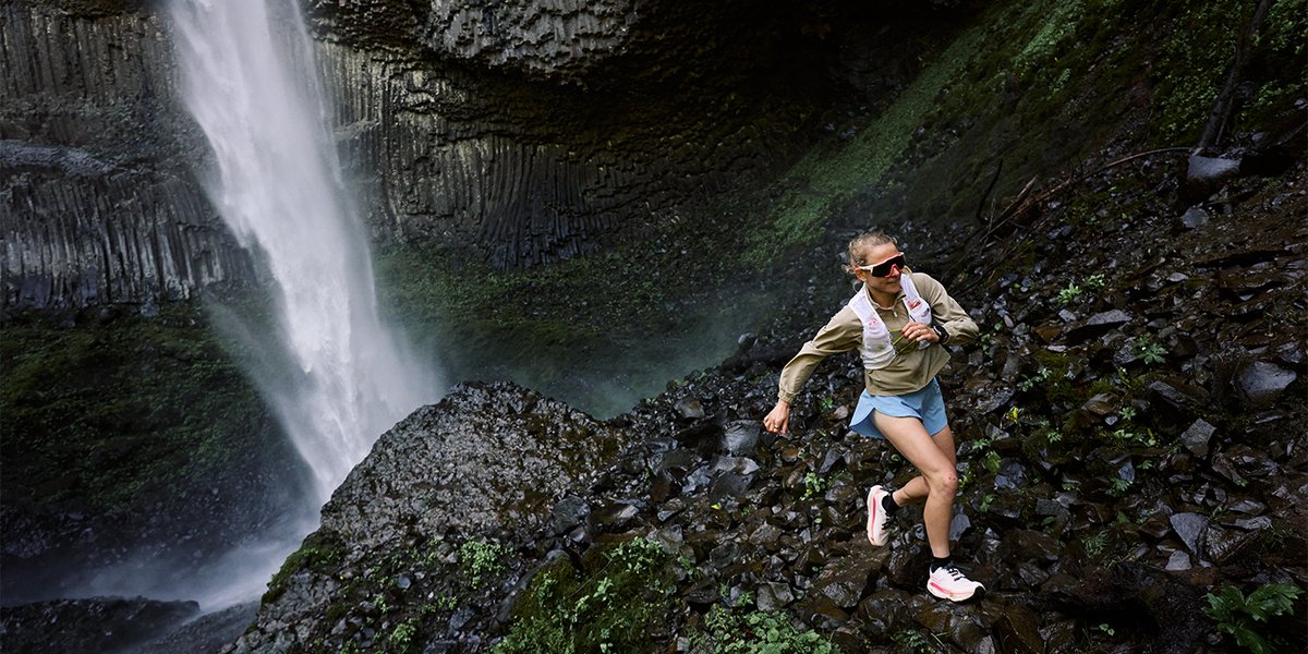 Person wearing Altra trail shoes sprinting down rocky slope in front of black rocks and dranatic waterfall in teh background.