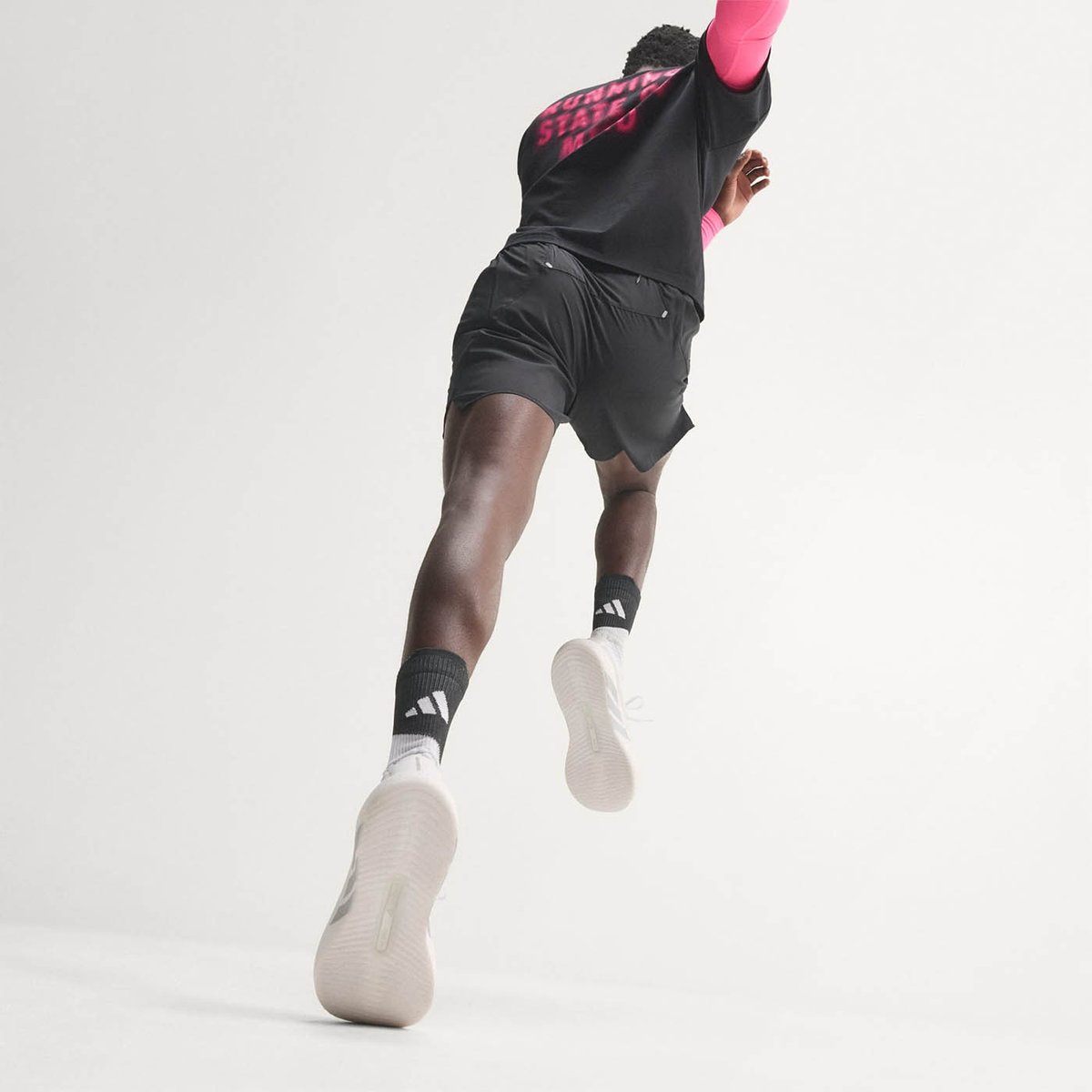 Runner in mid-stride from behind, wearing black shorts, black tee with pink graphic, pink arm sleeves, and white Adidas running shoes with off-white midsoles, on a white background.