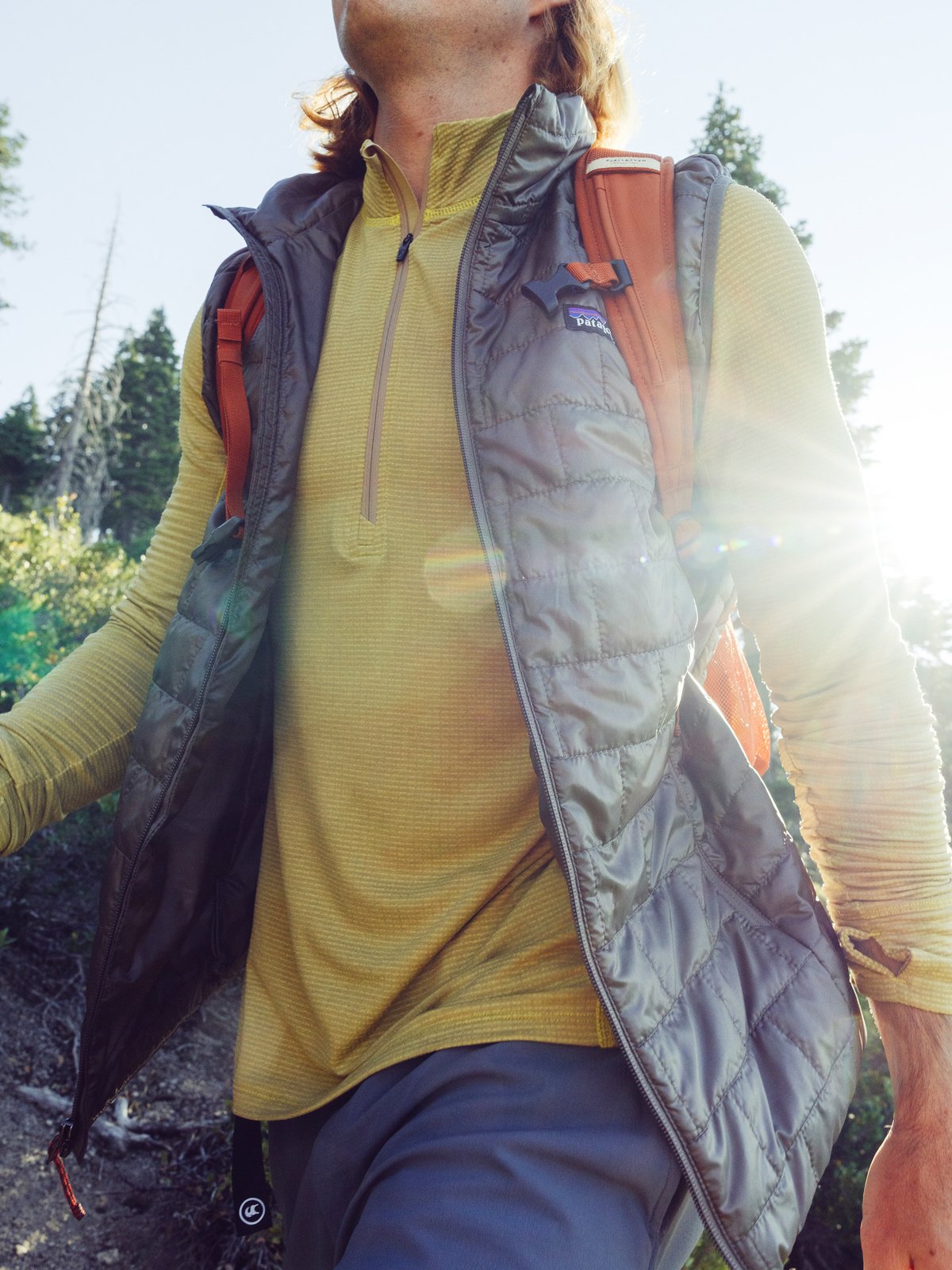 A person wearing a yellow long-sleeve shirt, a gray Patagonia vest, and an orange backpack hikes outdoors with sunlight behind him streaming through the trees.