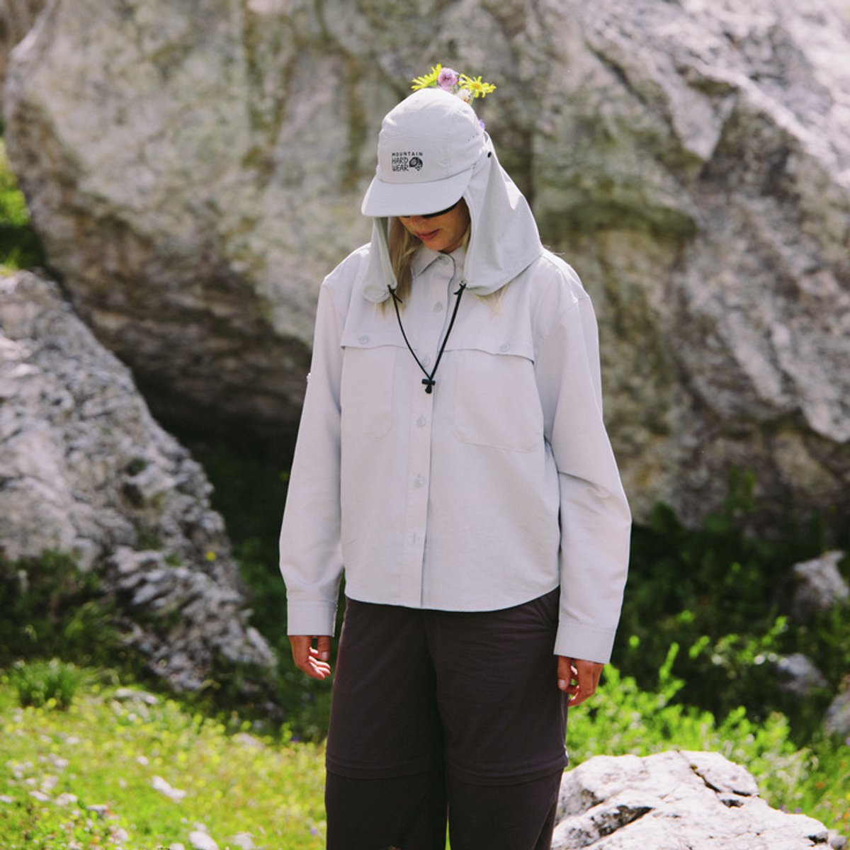 A woman stands outdoors, wearing a Sun Drift top with the hood on her head. A bouquet of flowers peeks out behind her hood.