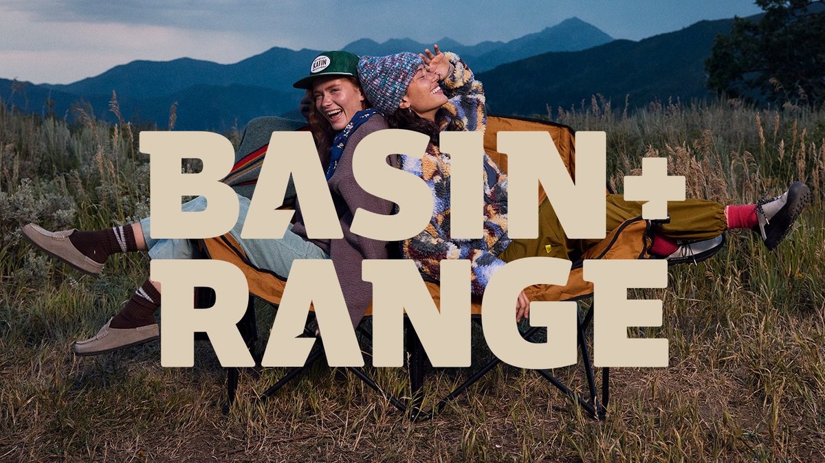 Two people laughing while lounging back-to-back in a camp chair in a grassy mountain meadow at dusk, wearing fleece and beanies, Basin + Range logo overlaid