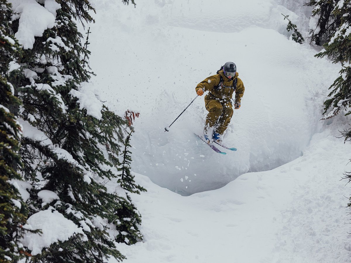Skier descending snowy pillow on tree covered hillside.