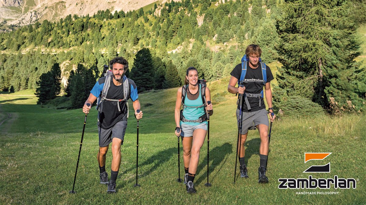 Three hikers trail running with trekking poles through an alpine valley with forested mountains in the background, showcasing outdoor adventure gear and active lifestyle.