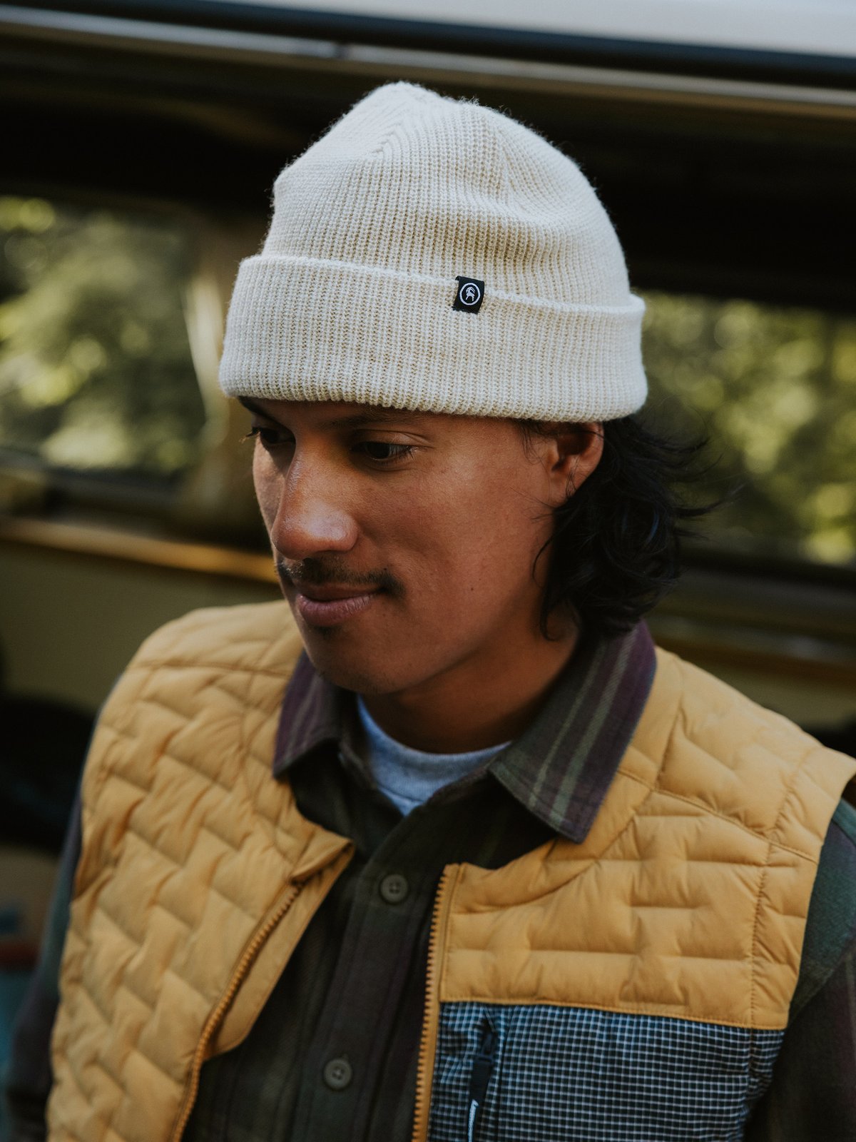 A man wearing a knit beanie and a quilted vest over a checkered button-up shirt, standing outdoors near a vehicle.